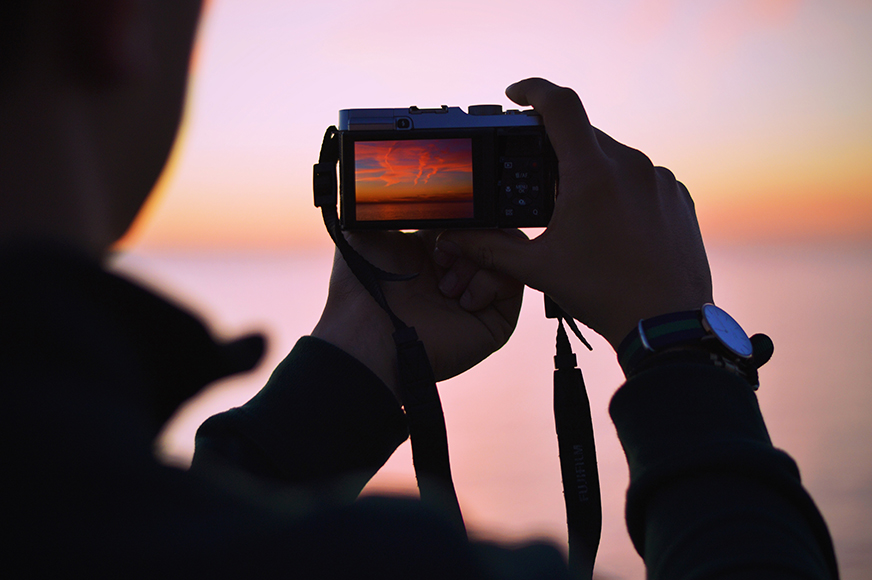 A person holding a camera takes a photo of a sunset over water, with the sunset reflected on the camera screen.
