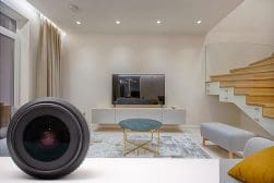 A modern living room with a large flat-screen TV on a white entertainment unit, a round coffee table with a green top, and a staircase with a wooden finish. A camera lens is in the foreground.