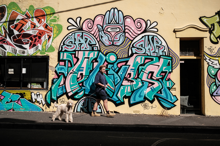 Man walking a dog past a colorful graffiti wall.