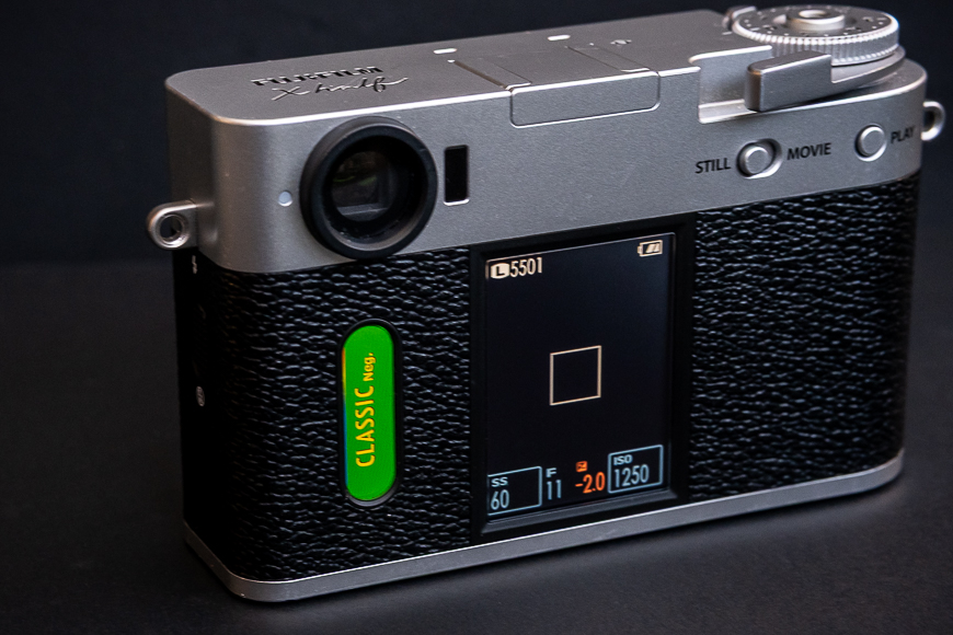 A digital camera with a textured black grip, silver body, and a display screen showing camera settings. The word "CLASSIC" appears on a green label beside the screen.