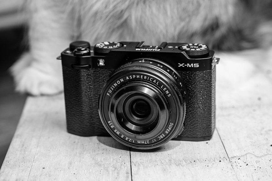 Black and white image of a Fujifilm X-M5 camera with a Fujinon Aspherical lens, placed on a wooden surface.