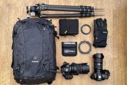 Photography gear laid out on a wooden floor, including a black backpack, two cameras, lenses, tripods, filters, and gloves.