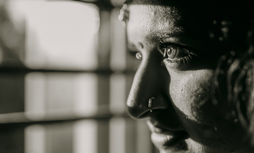 Close-up black and white photo of a person's face in profile, focusing on their eye and nose ring, with light coming through window bars in the background.