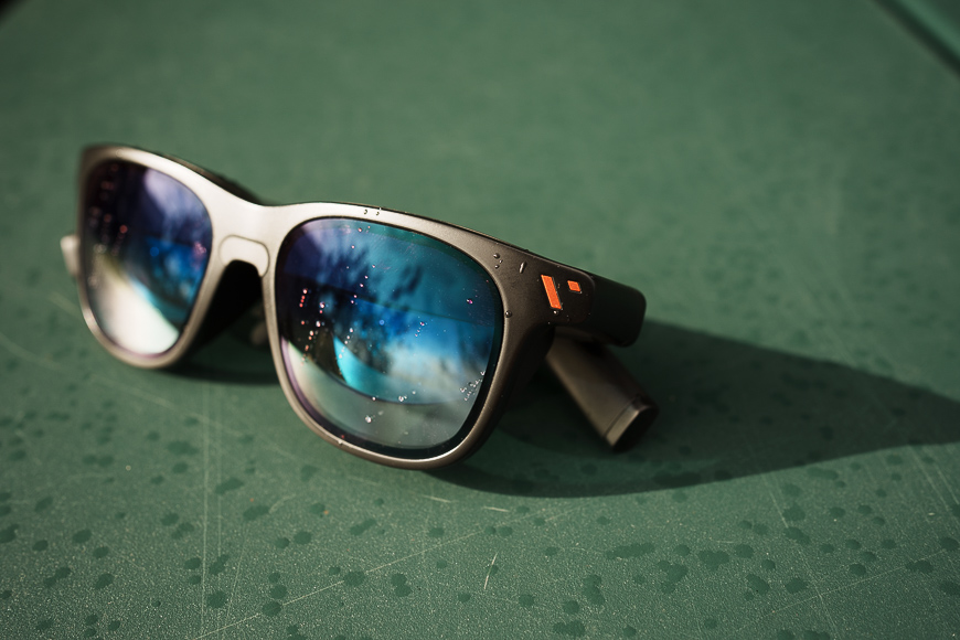 A pair of sunglasses with blue reflective lenses rests on a green surface with water droplets.