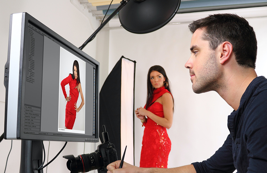 A photographer reviews a digital image of a woman in a red dress on a monitor while the model stands posing in a studio setup.