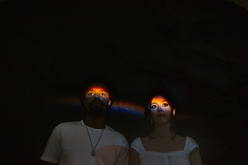 Two people in white shirts stand in the dark, illuminated by beams of light shining on their faces, highlighting their eyes.