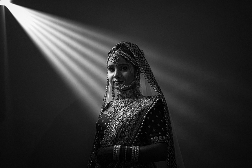 A woman in traditional bridal attire stands in partial darkness, illuminated by a narrow beam of light from the upper left, highlighting her ornate jewelry and clothing.