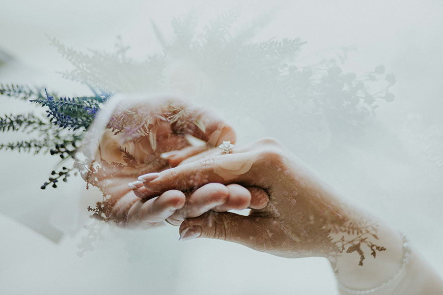 Close-up of two hands gently holding each other, with a soft overlay of flowers and foliage in the background. A ring is visible on one finger.