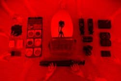 Flat lay of camera gear including a camera, lenses, filters, a tripod, a camera bag, and accessories, all arranged neatly on a textured red background. Multiple ghosted hands are visible.