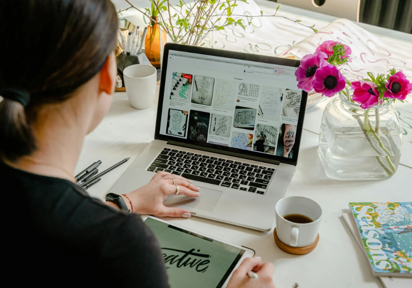 A person is working at a desk with a laptop displaying art images, a tablet, a cup of coffee, and flowers in a vase nearby. A person is working at a desk with a laptop displaying art images, a tablet, a cup of coffee, and flowers in a vase nearby.